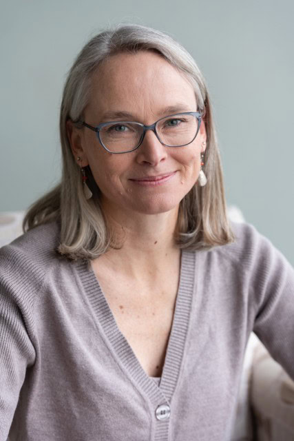 A woman with gray hair and glasses, wearing a beige cardigan, is sitting and smiling at the camera in front of a light green background. She is one of our upcoming guests.