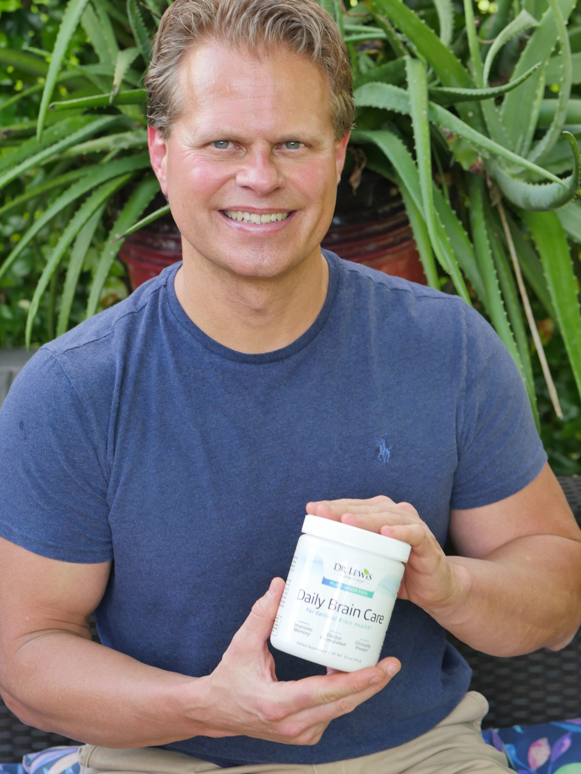 Dr. John Lewis sits outdoors, smiling, and holding a container labeled "Daily Brain Care," embodying wellness. Aloe plants thrive in the background, creating a serene setting for both body and mind.