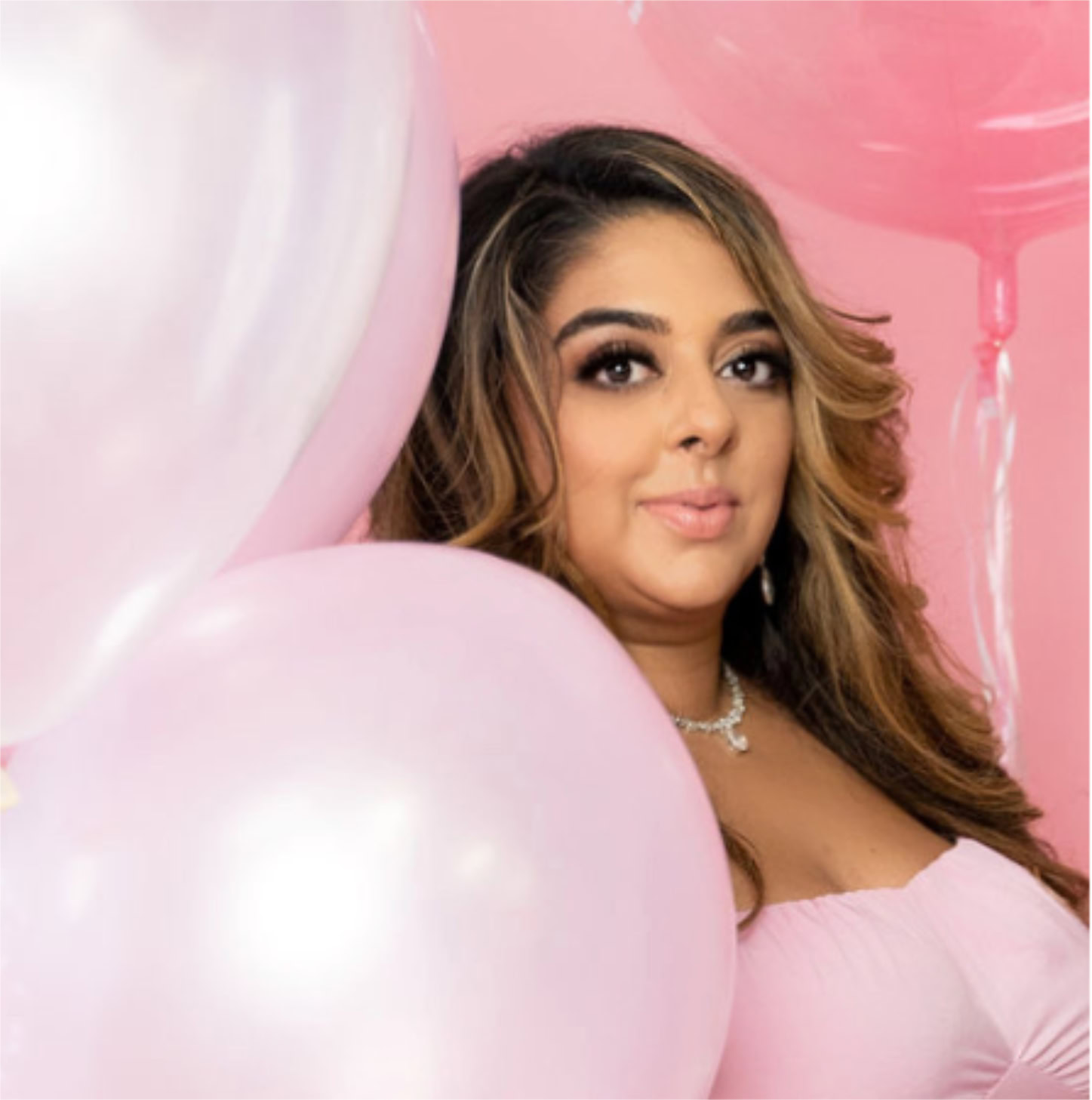 A woman with long hair stands next to pink and white balloons against a pink background, wearing a stylish pink top and necklace, setting the stage for upcoming guests.