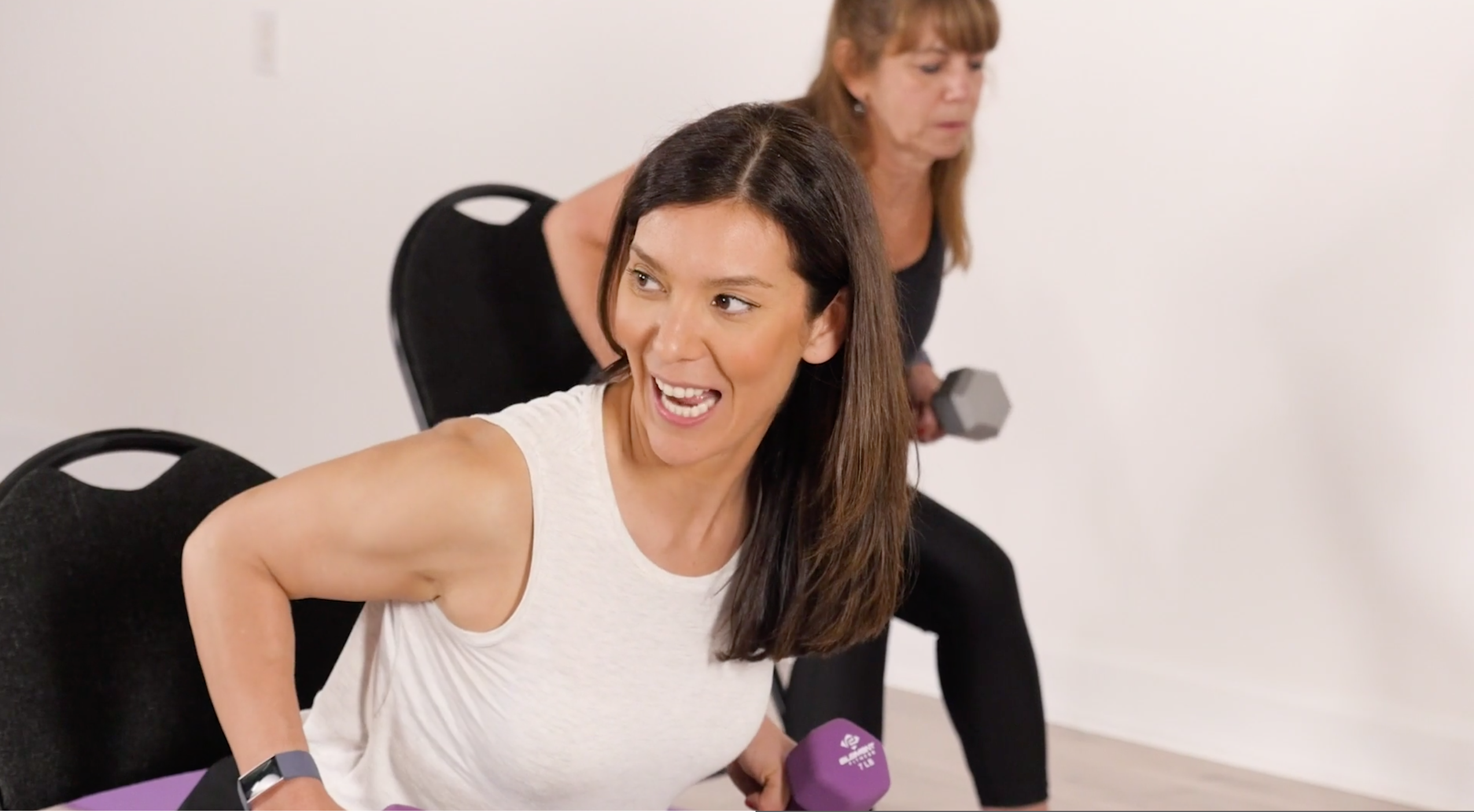 Two women, embracing strength over 50, exercise with dumbbells while seated on black chairs in a bright room.