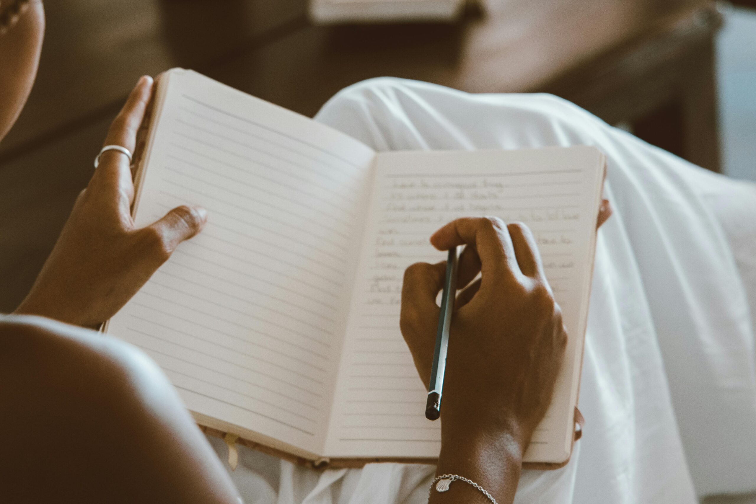 In a white garment, a person pens down their aspirations for the best year yet in 2025, writing thoughtfully in a lined notebook.