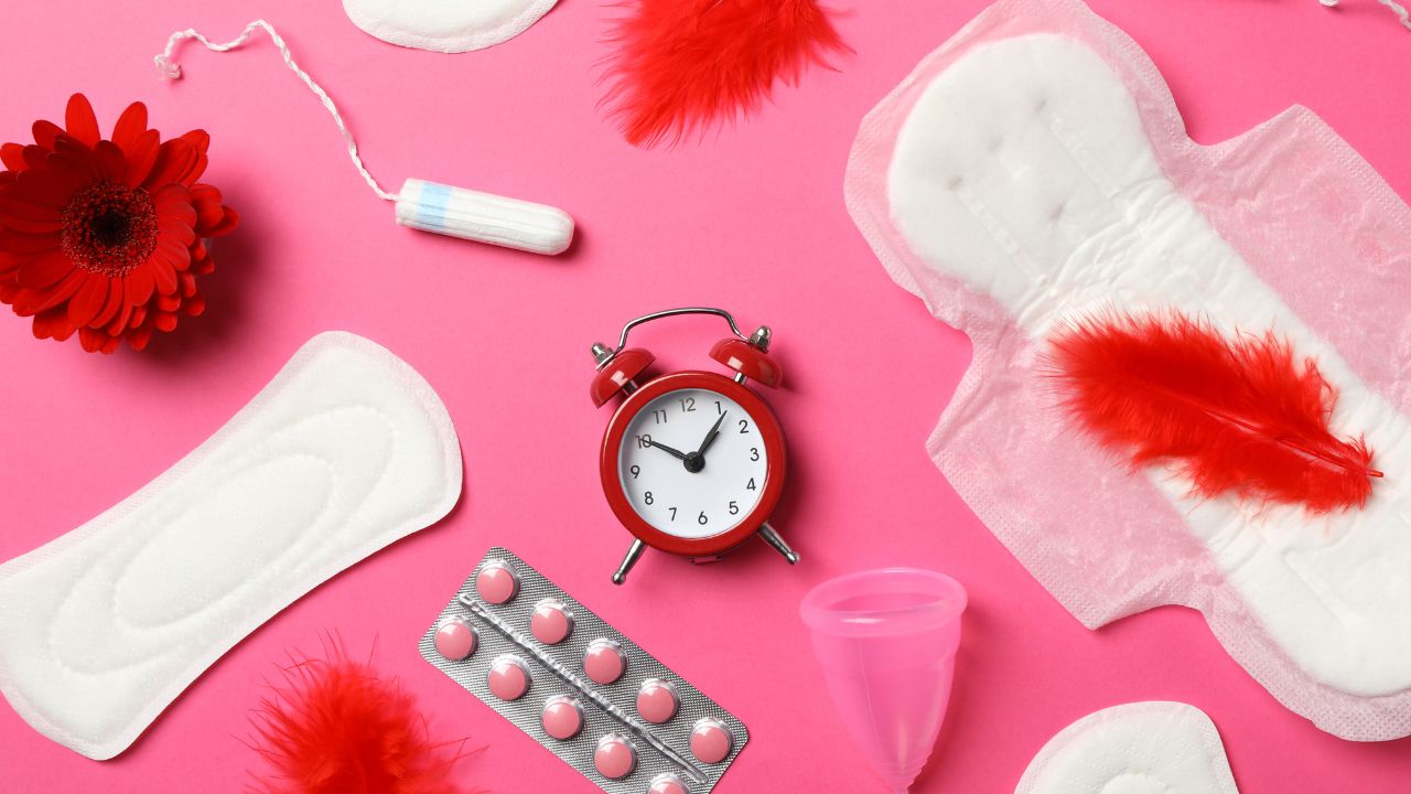 Menstrual products, a red alarm clock, red feathers, and a red flower are arranged on a pink background, symbolizing the urgency and importance of women's health. Cervical Health Can't Wait reminds us to prioritize these vital conversations.