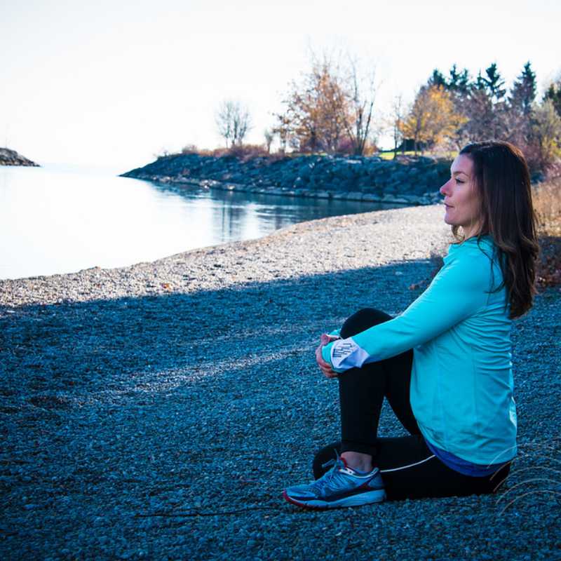 A woman with surprising strength sits on a pebble beach by a calm lake, wearing a blue jacket and black leggings, with trees and a rocky shoreline in the background.