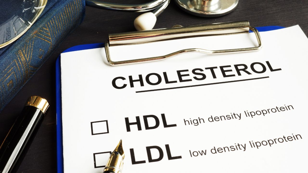 A clipboard displays text about cholesterol with checkboxes for HDL (high-density lipoprotein) and LDL (low-density lipoprotein), alongside a pen and stethoscope, highlighting heart health risks.