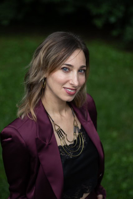 Evalee Gertz of Sia Silk with shoulder-length hair, wearing a maroon jacket and black top, stands outdoors amidst lush greenery, looking as if they've just awoken from deep sleep.