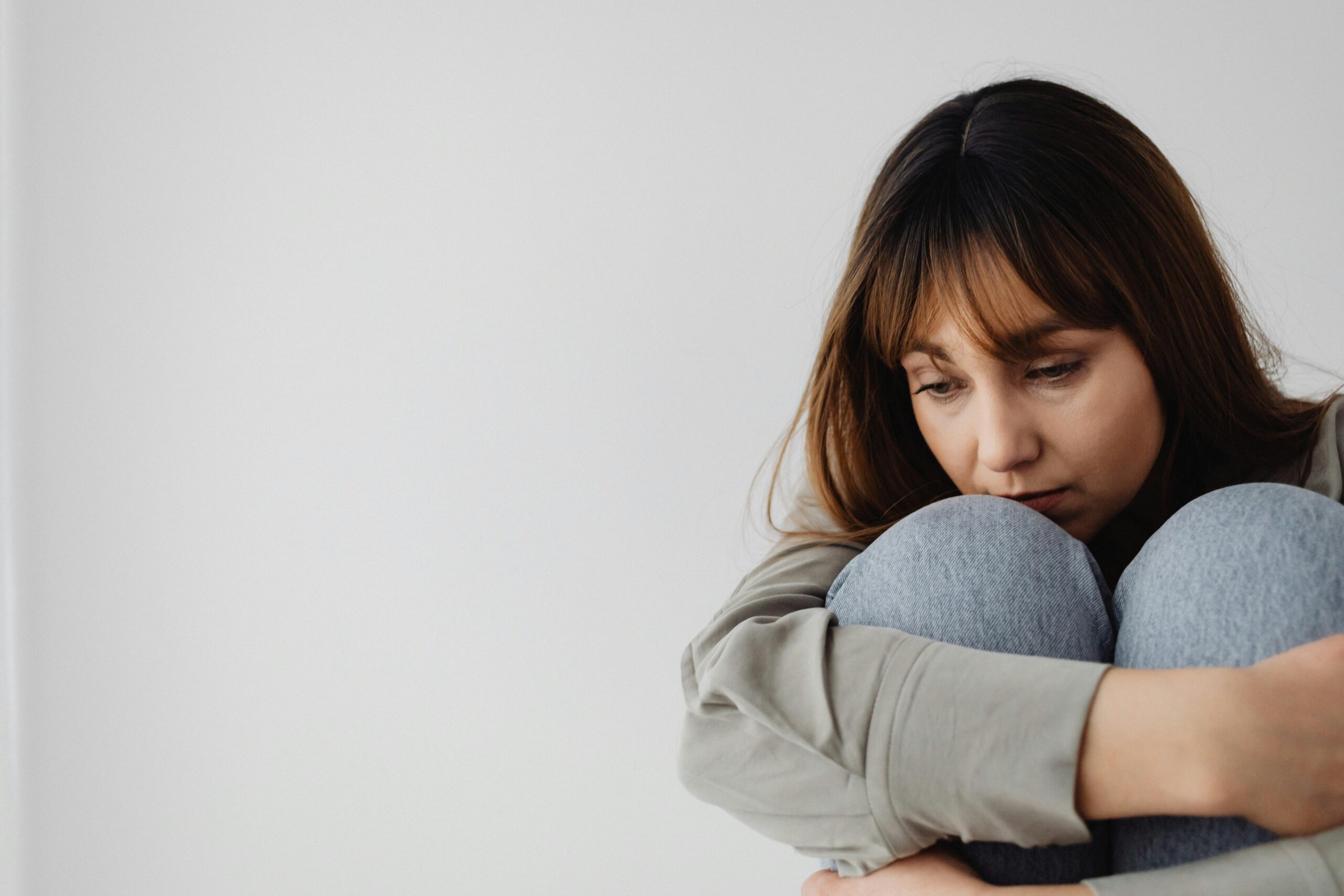 A woman with long brown hair sits against a white wall, hugging her knees and looking down with a pensive expression, seeking relief from menopausal depression.