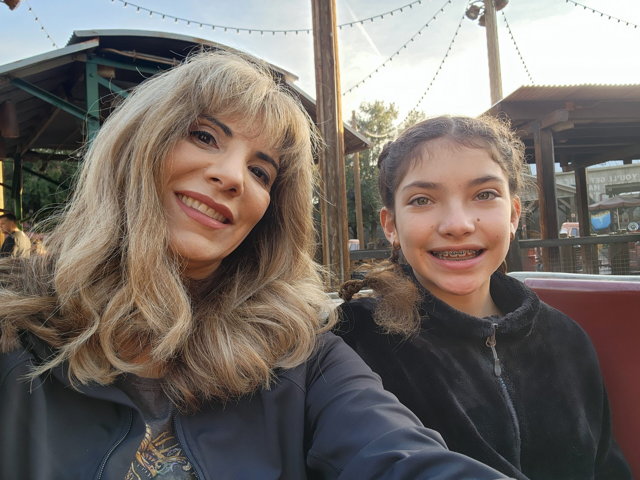 Carmen and Jasmine Hecox smiling for a selfie beneath enchanting string lights and rustic wooden structures, embracing the joy of the moment as they celebrate finding relief from menopause.