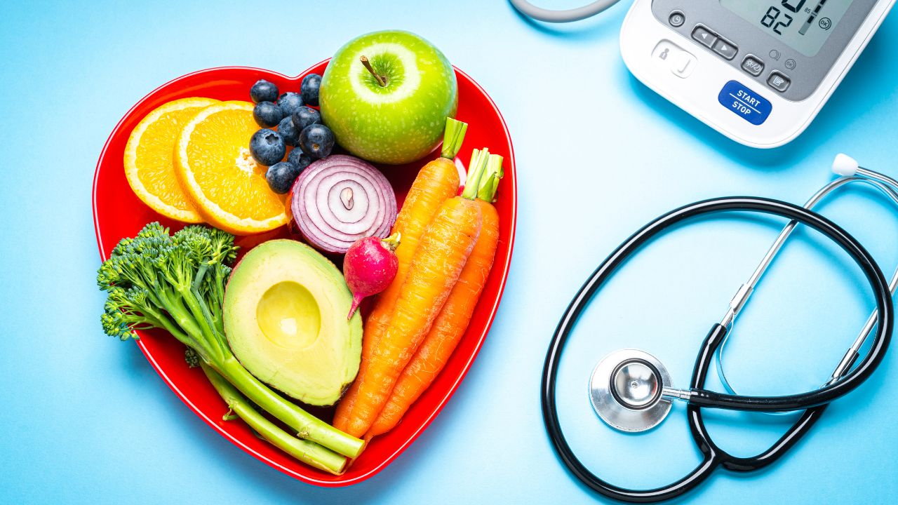 A red heart-shaped bowl with fruits and vegetables sits next to a blood pressure monitor and a stethoscope on a blue surface, highlighting the importance of heart health risks in menopause.