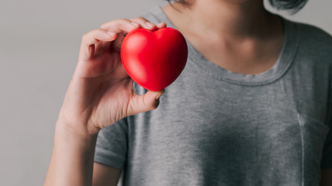 A person in a gray shirt holds a red heart-shaped object close to their chest, symbolizing the importance of heart health and awareness, especially pertinent for women as they navigate menopause and the unique risks of women's heart attacks.