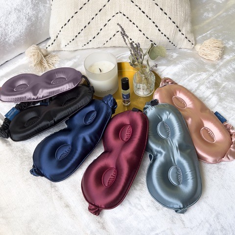 A collection of six Sia Silk satin eye masks in various colors rests on a bed, promising quality sleep. Nearby, a lit candle, a small vase with dried flowers, and a roll-on bottle create a serene atmosphere.