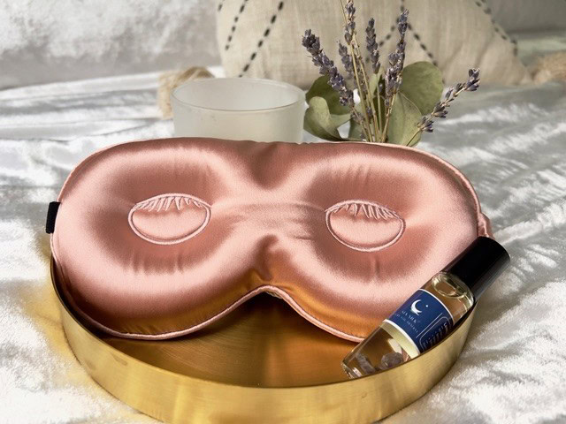 Silk sleep mask and Sia Silk essential oil roller rest on a gold tray, placed on white fabric, with a candle and lavender sprigs in the background—all crafted for deep restorative sleep, inspired by Evalee Gertz's serene design.