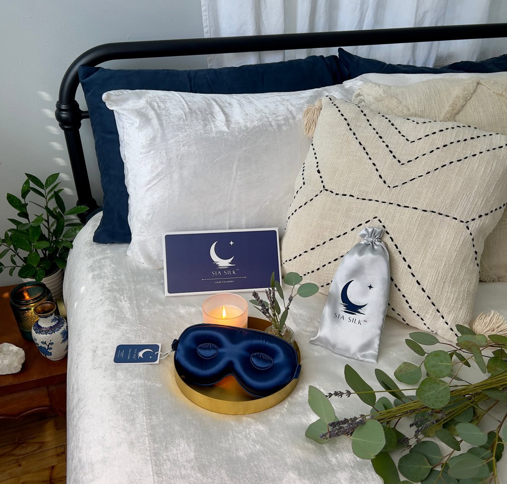 A tranquil bedroom scene features a bed adorned with blue and white pillows, a soothing blue eye mask for better rest, and a gentle candle on a tray. A Sia Silk pouch sits elegantly nearby, while lush plants add life. The arrangement is completed by an intriguing box with a moon design.