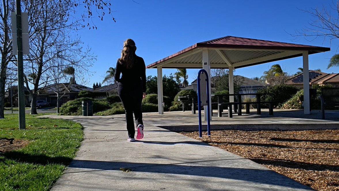 A person, embracing the power of outdoor activity, strolls along a path in the park toward a pavilion with picnic tables, surrounded by trees and houses beneath the clear blue sky.