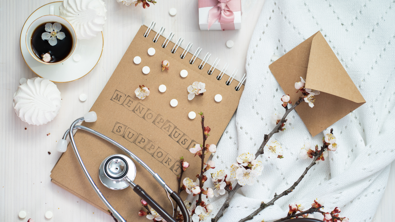 A notepad with "Menopause Support" text sits amid pills, a stethoscope, coffee cup, pink gift box, flowers, and an envelope on a light surface—highlighting the essential care during perimenopause.