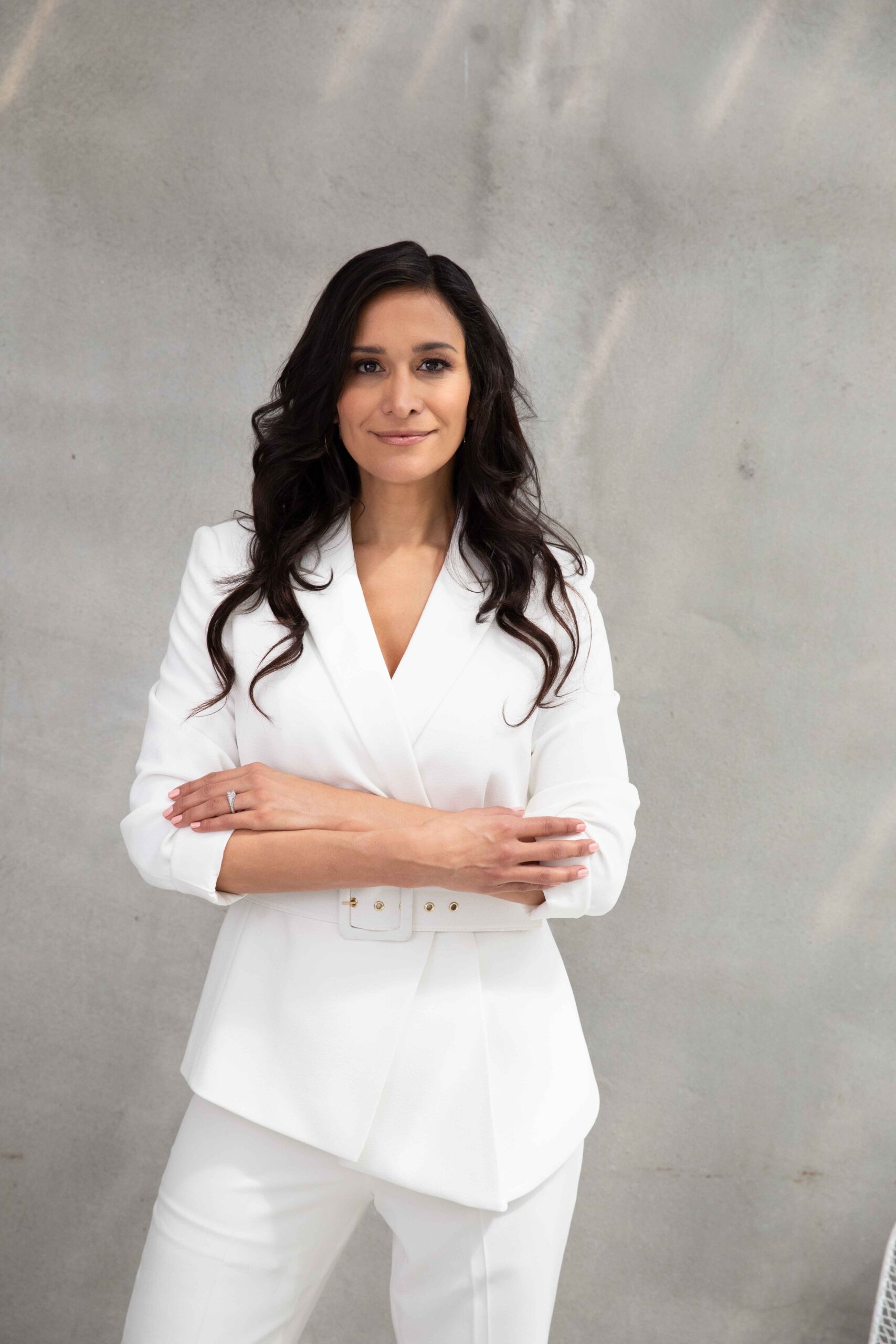 Carin Luna-Ostaseski with long dark hair in a white suit stands against a light gray background, arms crossed, portraying confidence and empowerment as she faces forward. She embodies the strength needed for debunking menopause myths: it’s more than just hot flashes.