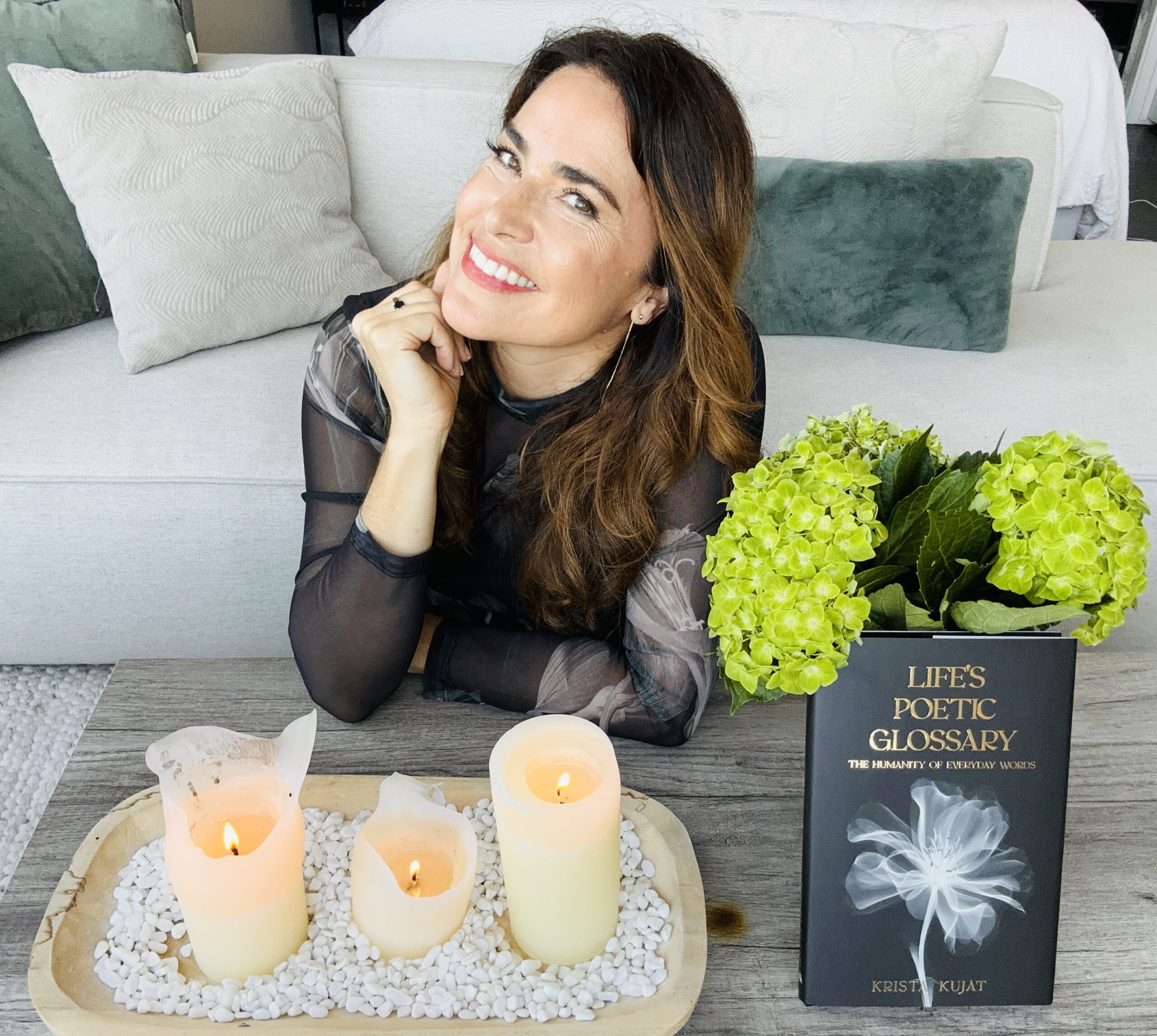 Rhiah Kujat sits at a table with three lit candles on a tray of white stones, a vase of green hydrangeas, and a book titled "Life's Poetic Glossary," reflecting on turning betrayal into empowerment.