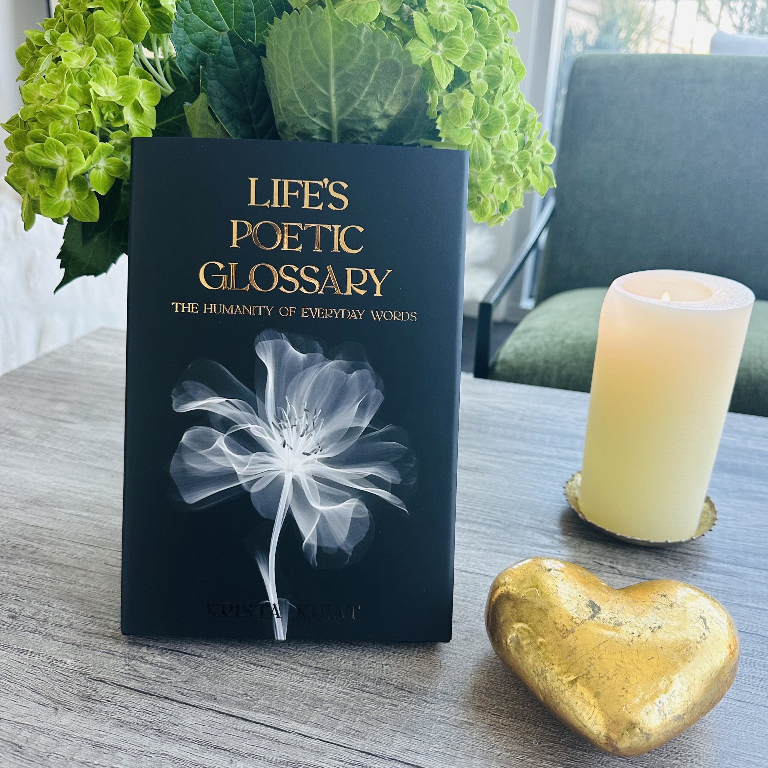 A book titled "Life's Poetic Glossary" stands on a table beside a lit candle, a gold heart decoration, and green hydrangeas—inviting you to explore understanding the power of somatic healing in everyday moments.