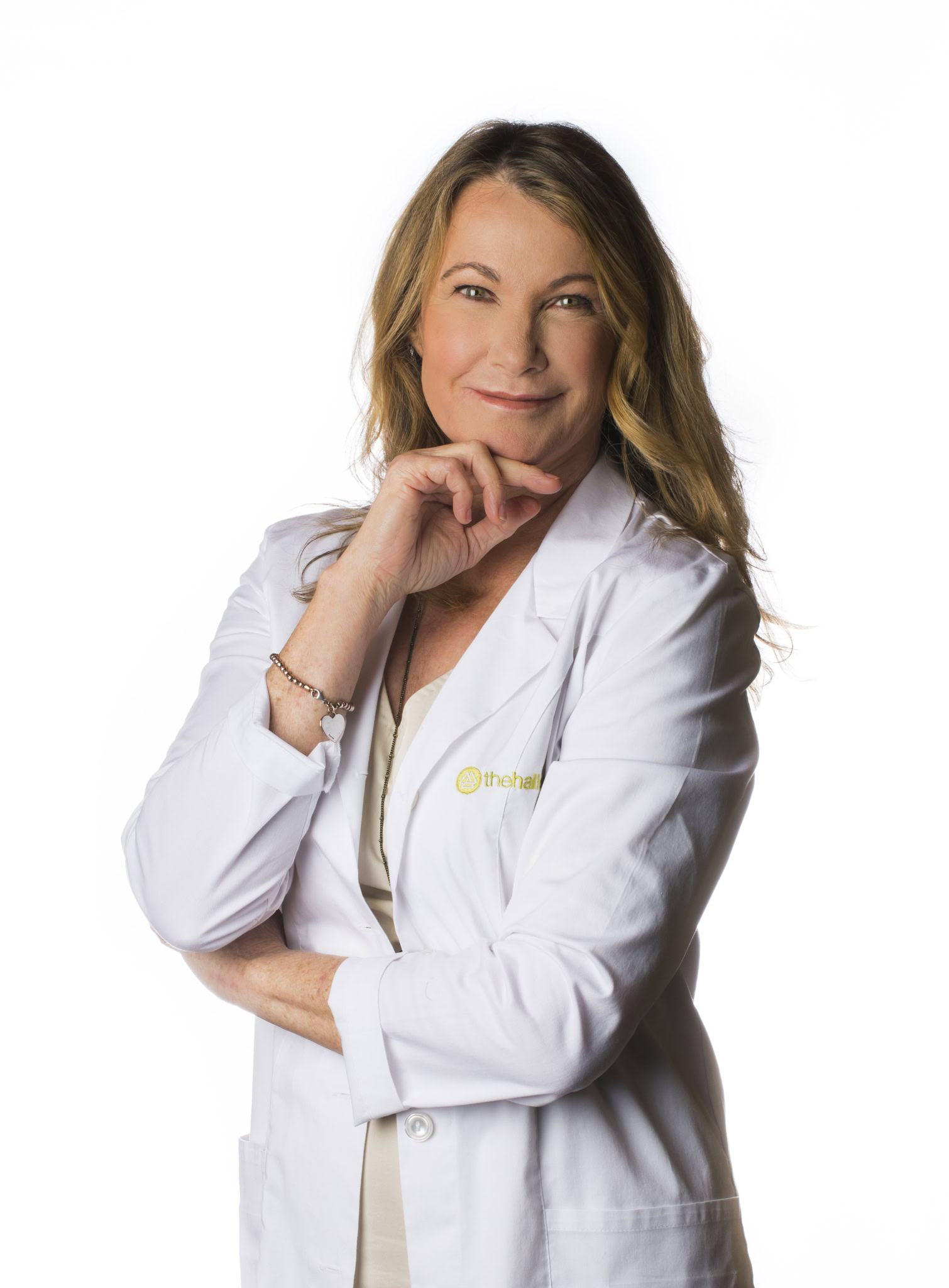 Upcoming Guest's A woman with long blonde hair wearing a white lab coat stands against a plain white background, smiling with her hand resting on her chin, embodying women’s empowerment and confidence.