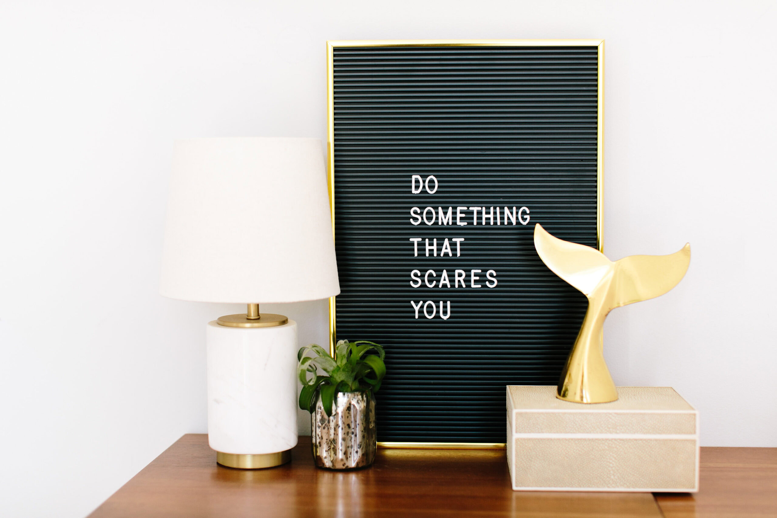 A letterboard with the phrase "DO SOMETHING THAT SCARES YOU" stands on a wooden surface beside a lamp, a small plant, and a gold whale tail ornament, inspiring you to start cultivating a growth mindset every day.