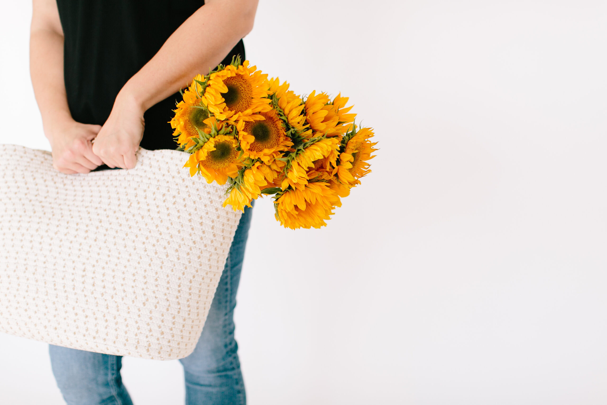 A person holding a woven white tote bag with a bouquet of yellow sunflowers stands against a plain white background, capturing the spirit of midlife and menopause: a shared yet unique journey full of new beginnings.