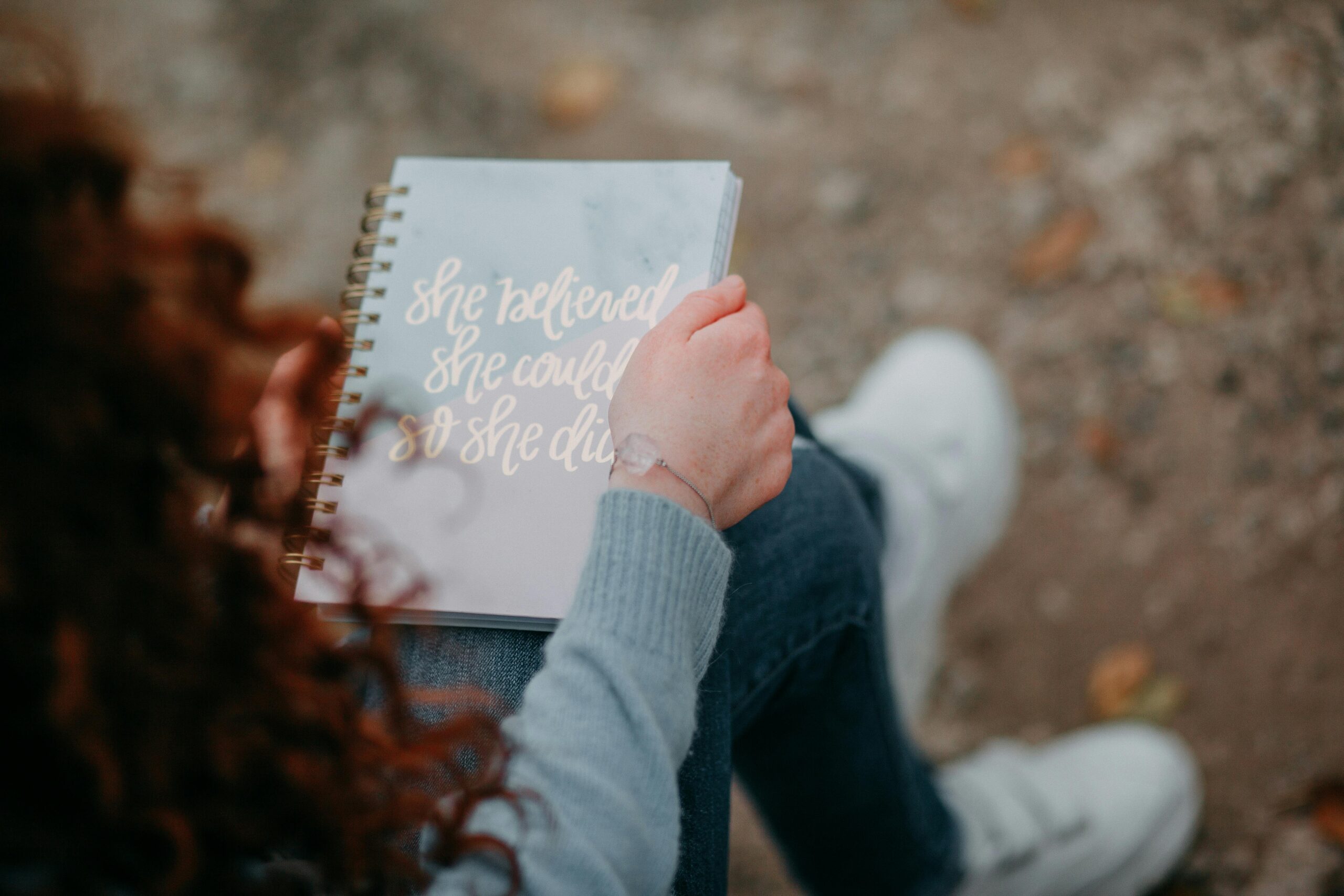 Person holding a notebook with the cover text "she believed she could so she did," sitting outdoors with their legs crossed, reflecting on Practical Steps to Uncover Your Midlife Purpose.