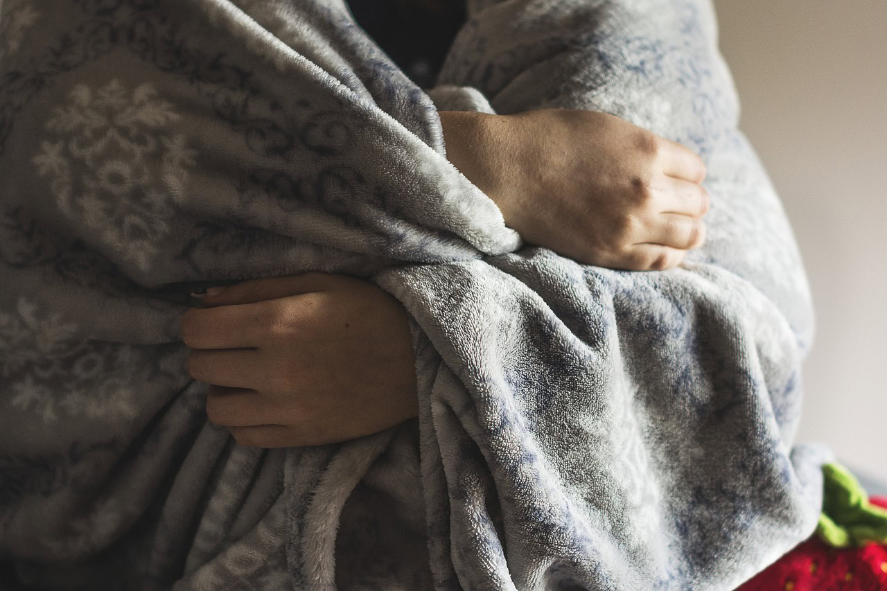 A person wraps themselves in a gray, patterned blanket, with their arms crossed over their chest—an image reminiscent of the real-life consequences found in mold-related health stories.