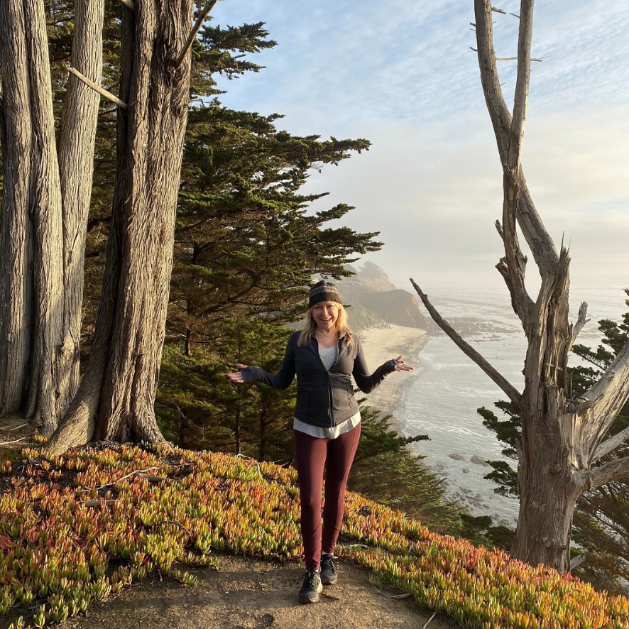 Erika Shershun, MA, MFT, stands on a coastal cliffside trail, surrounded by tall trees and plants, contemplating why traditional therapy might not be enough, with the ocean and coastline stretching out in the background.