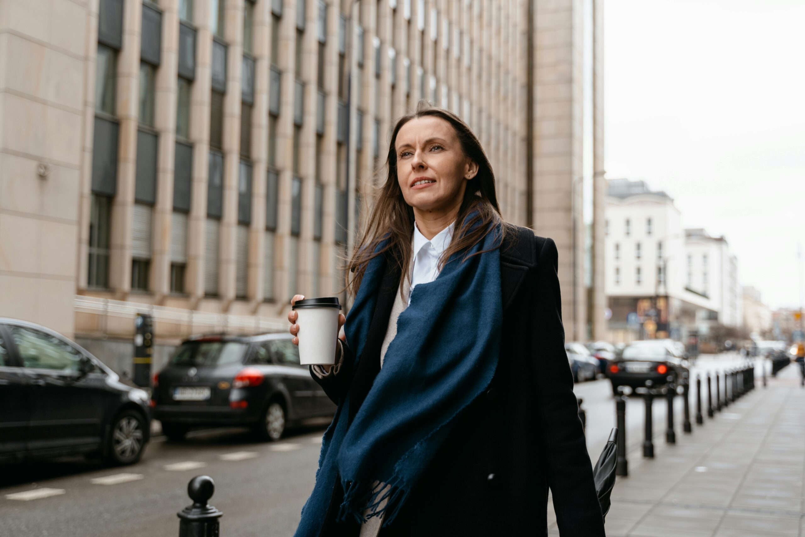 A woman in a dark coat and blue scarf walks down a city sidewalk holding a takeaway coffee cup, seamlessly integrating new habits into her daily routine amid cars and tall buildings in the background.