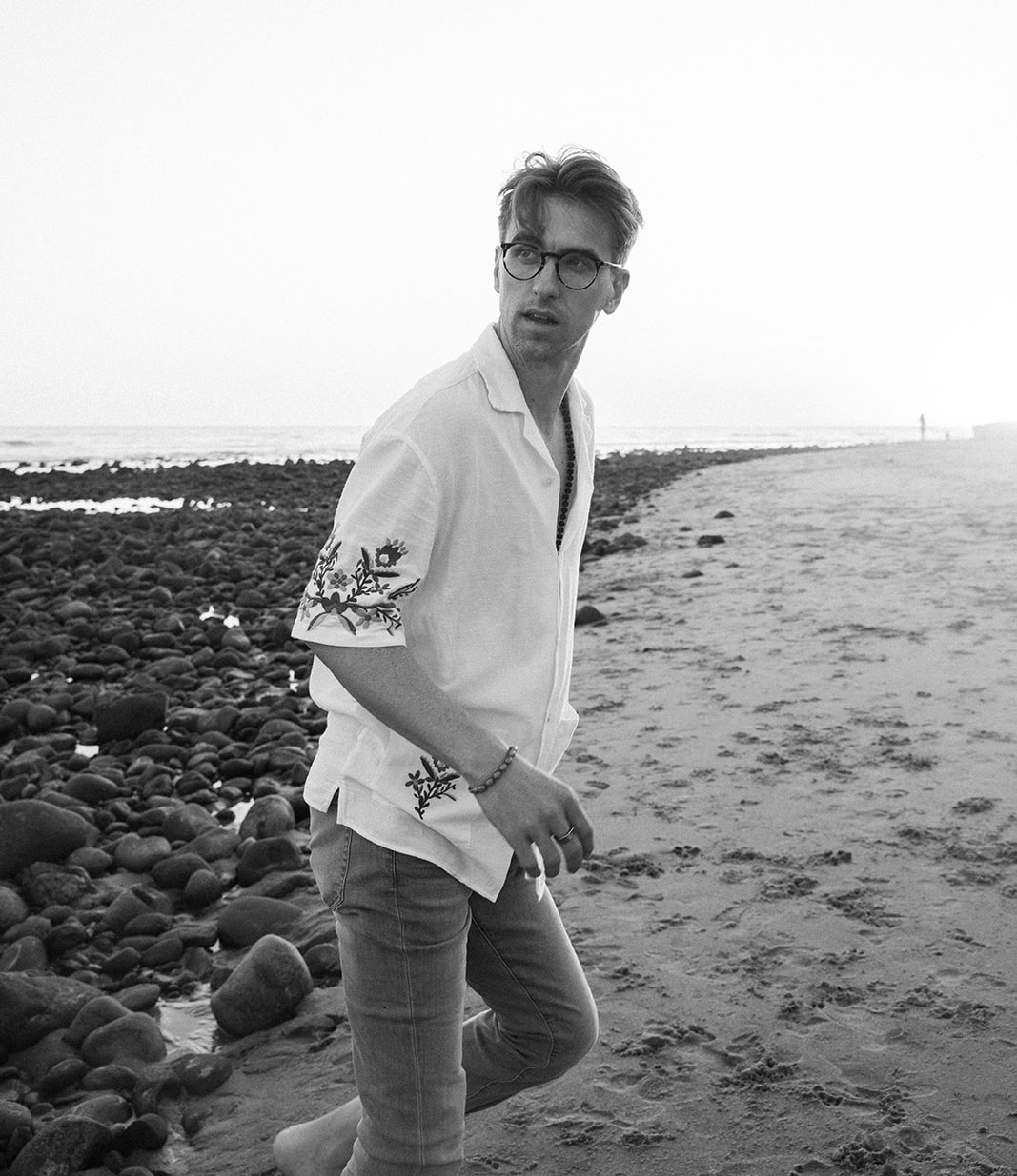 A man wearing glasses, a white shirt with floral embroidery, and jeans stands barefoot on a rocky beach, reflecting on how the perfect home shouldn't be making you sick.