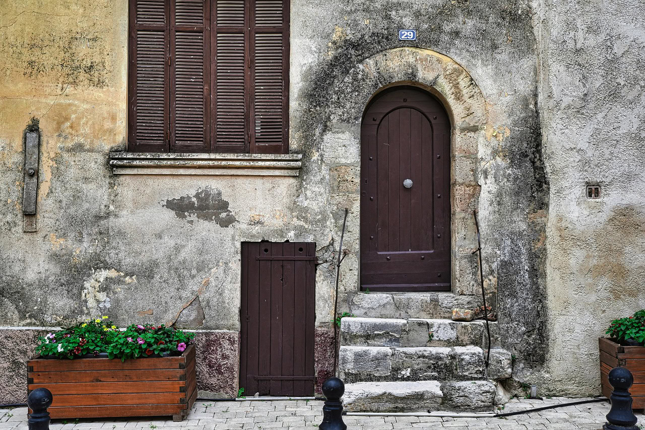 An old stone building with a brown arched door, small stairs, a shuttered window, and two wooden planters with flowers stands weathered by time—its walls quietly hinting at the silent threat of mold and mycotoxins within.