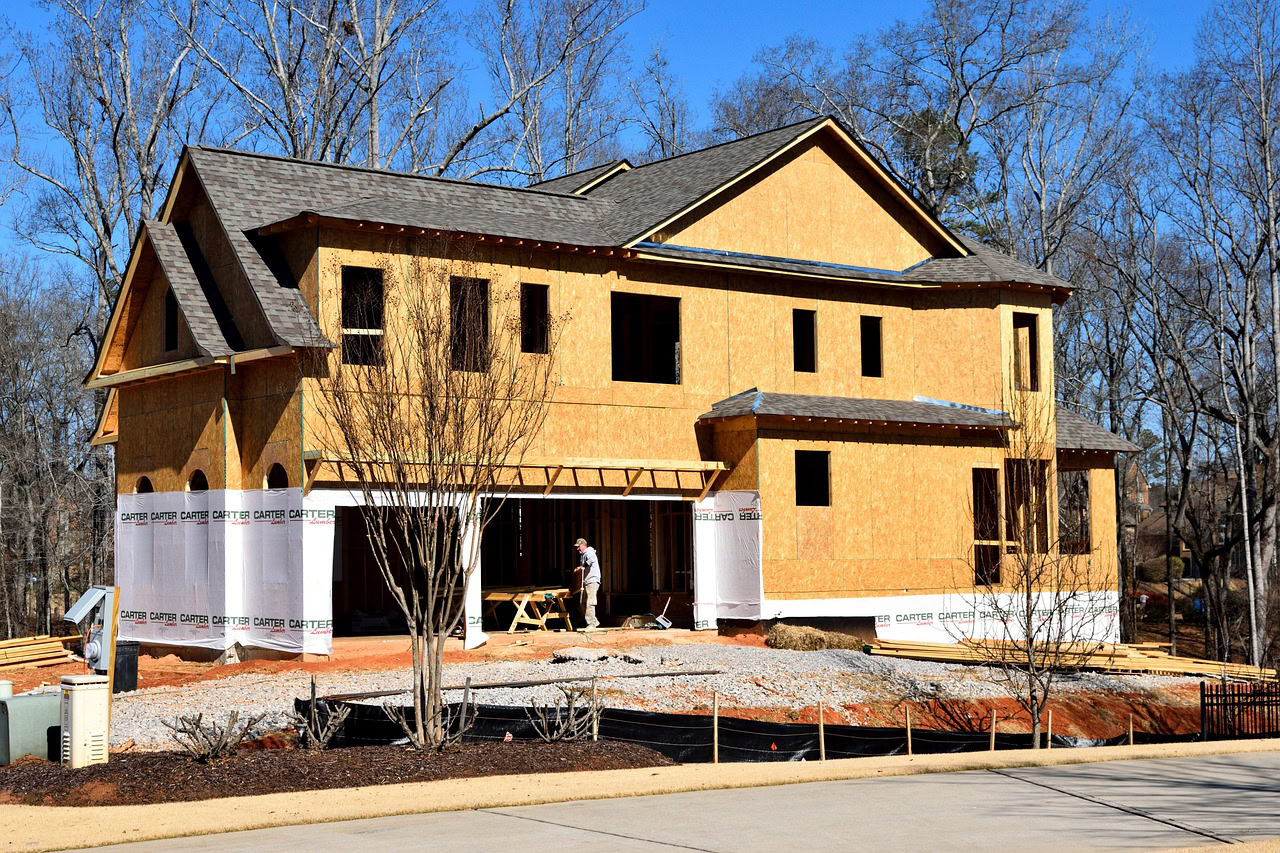 A two-story house under construction with plywood walls, exposed framing, and a worker standing by a table in the front yard highlights why modern homes are especially vulnerable during the building process.