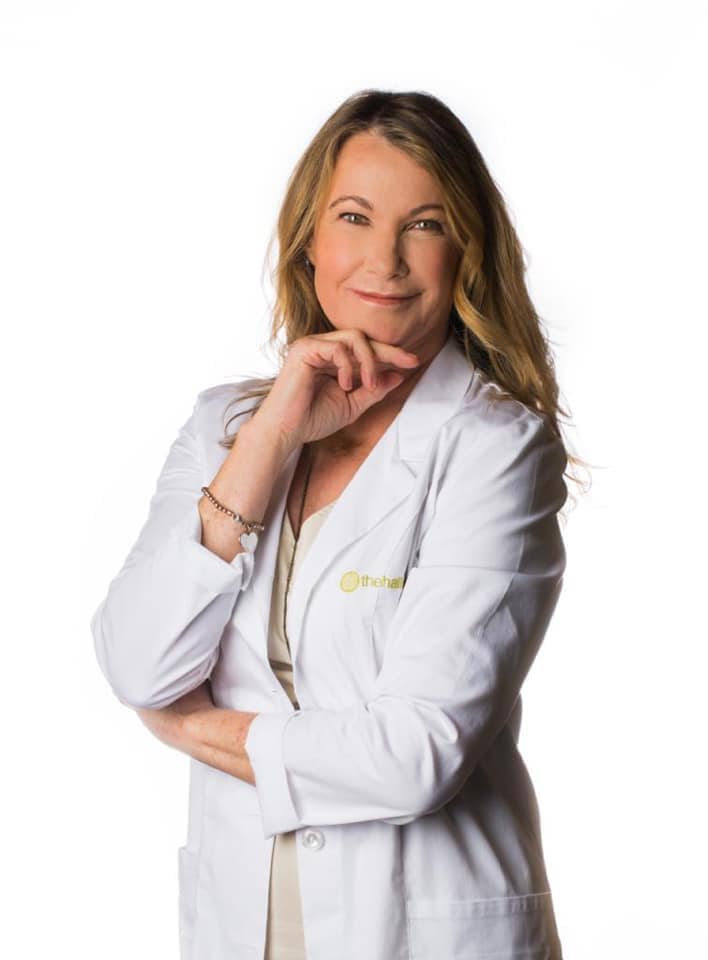 Dr. Prudence Hall in a white lab coat stands with one hand on her chin and the other arm crossed, posing confidently against a plain white background—symbolizing expertise in women's health and hormones.