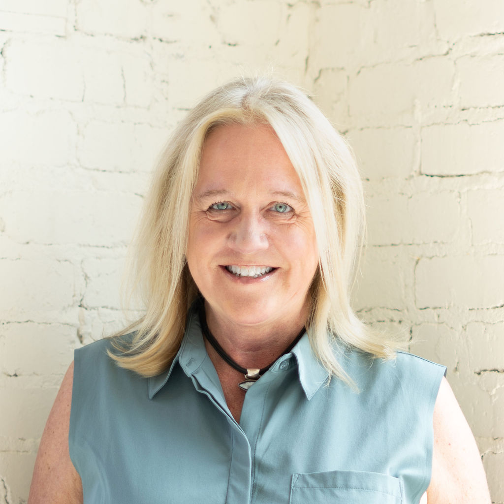 A woman with shoulder-length blonde hair, wearing a sleeveless light blue top and a black necklace, smiles in front of a white brick wall—an inspiring presence among our Upcoming Guests.