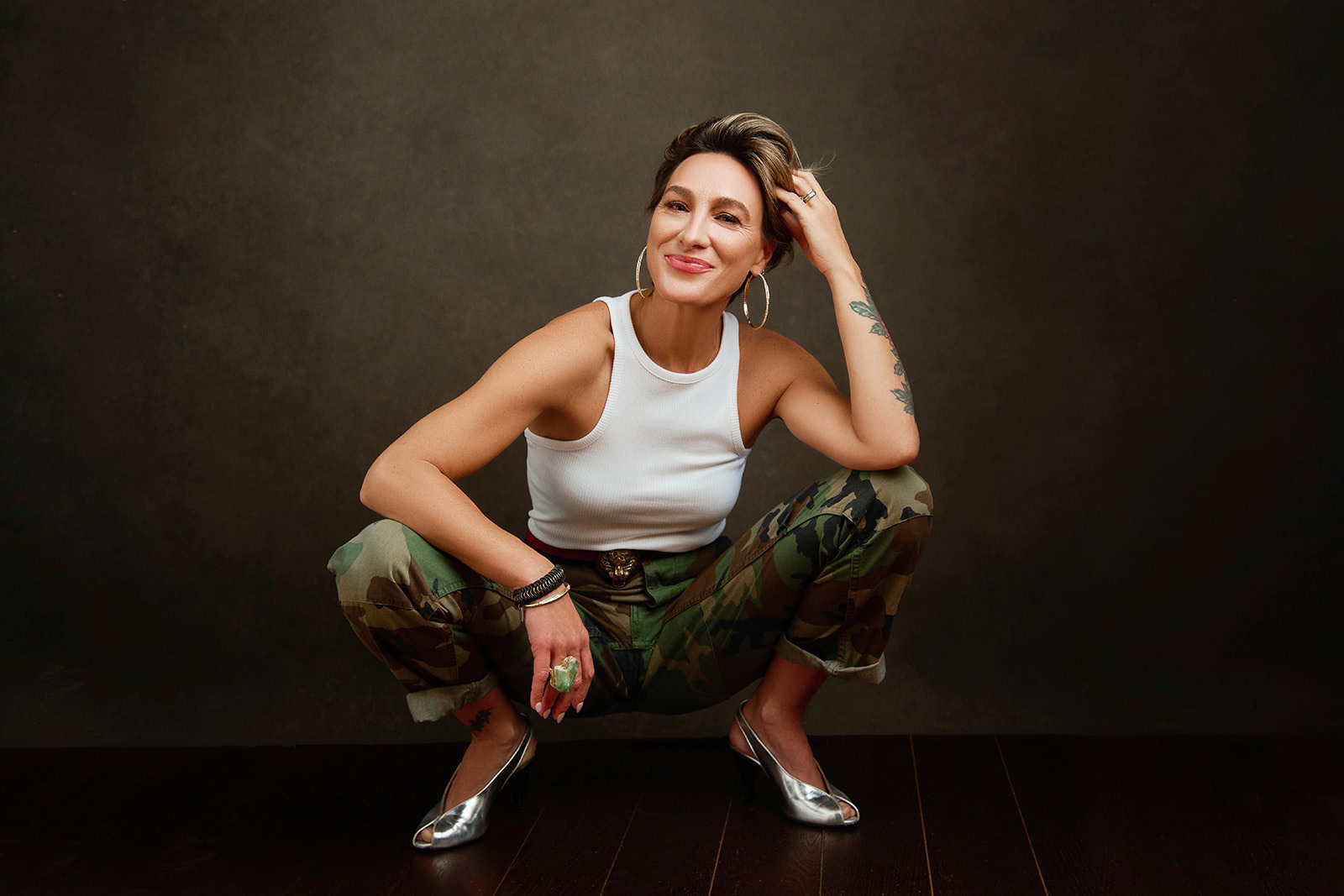 Natty Frasca with short hair squats, smiling, wearing a white sleeveless top, camouflage pants, hoop earrings, bracelets, and silver heels against a dark backdrop—embodying overcoming fear and embracing change with confidence.