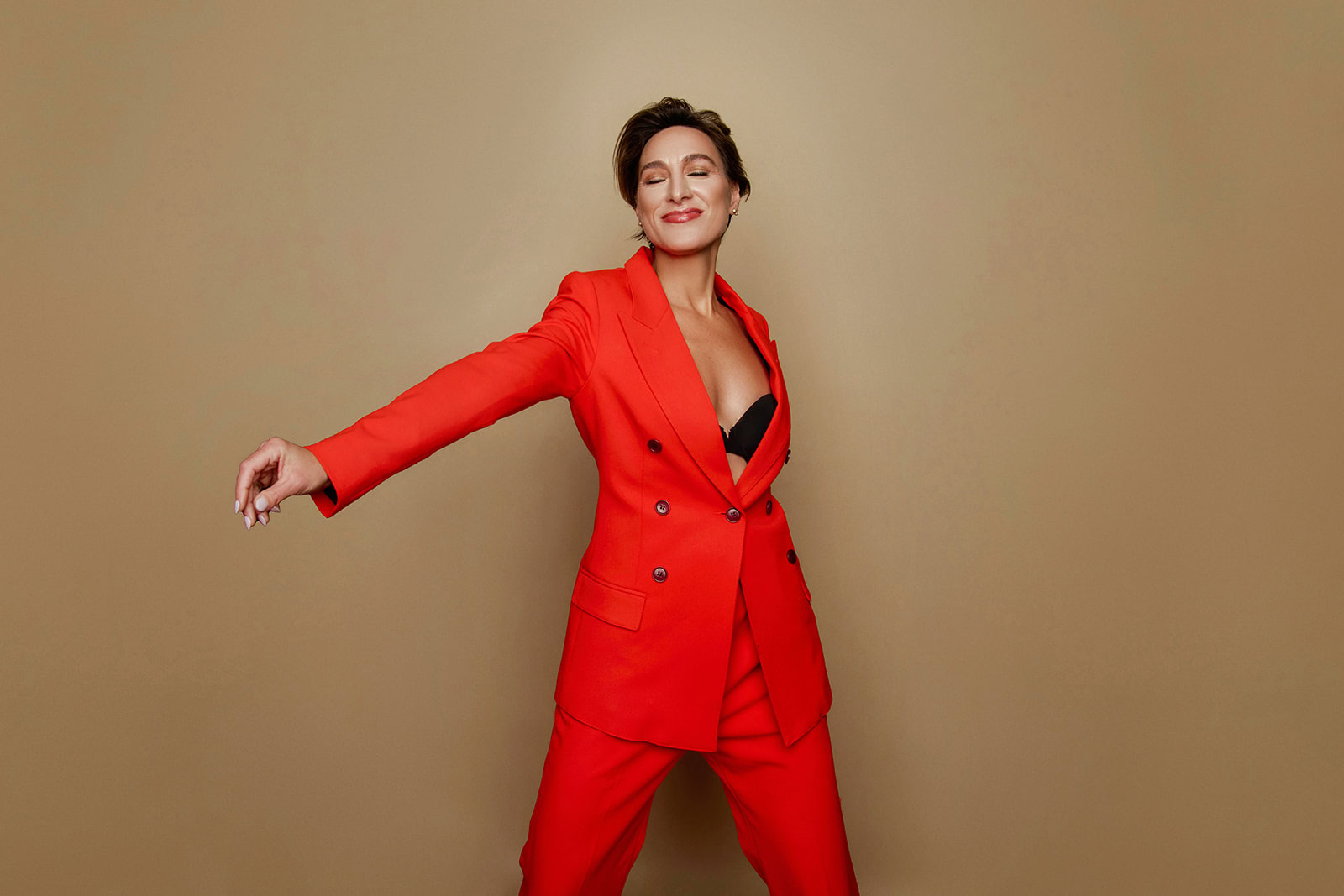 A person wearing a bright red suit and black top poses confidently against a plain beige background with one arm extended, capturing the essence of rediscovering pleasure in midlife—much like Natty Frasca’s spirited journey.